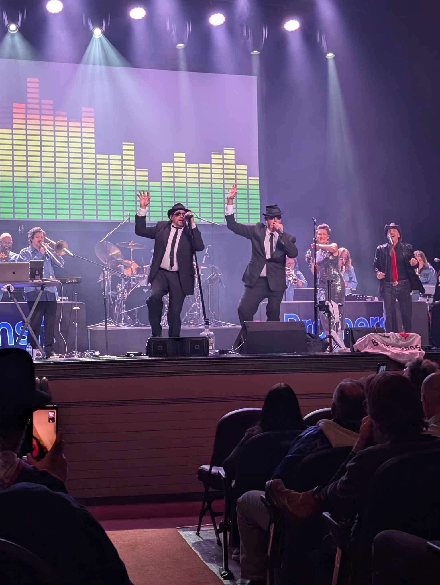 Wide theater-performance view of the Transit Brothers Blues Brothers Experience showing the full stage, band, featured performers, lighting and audience